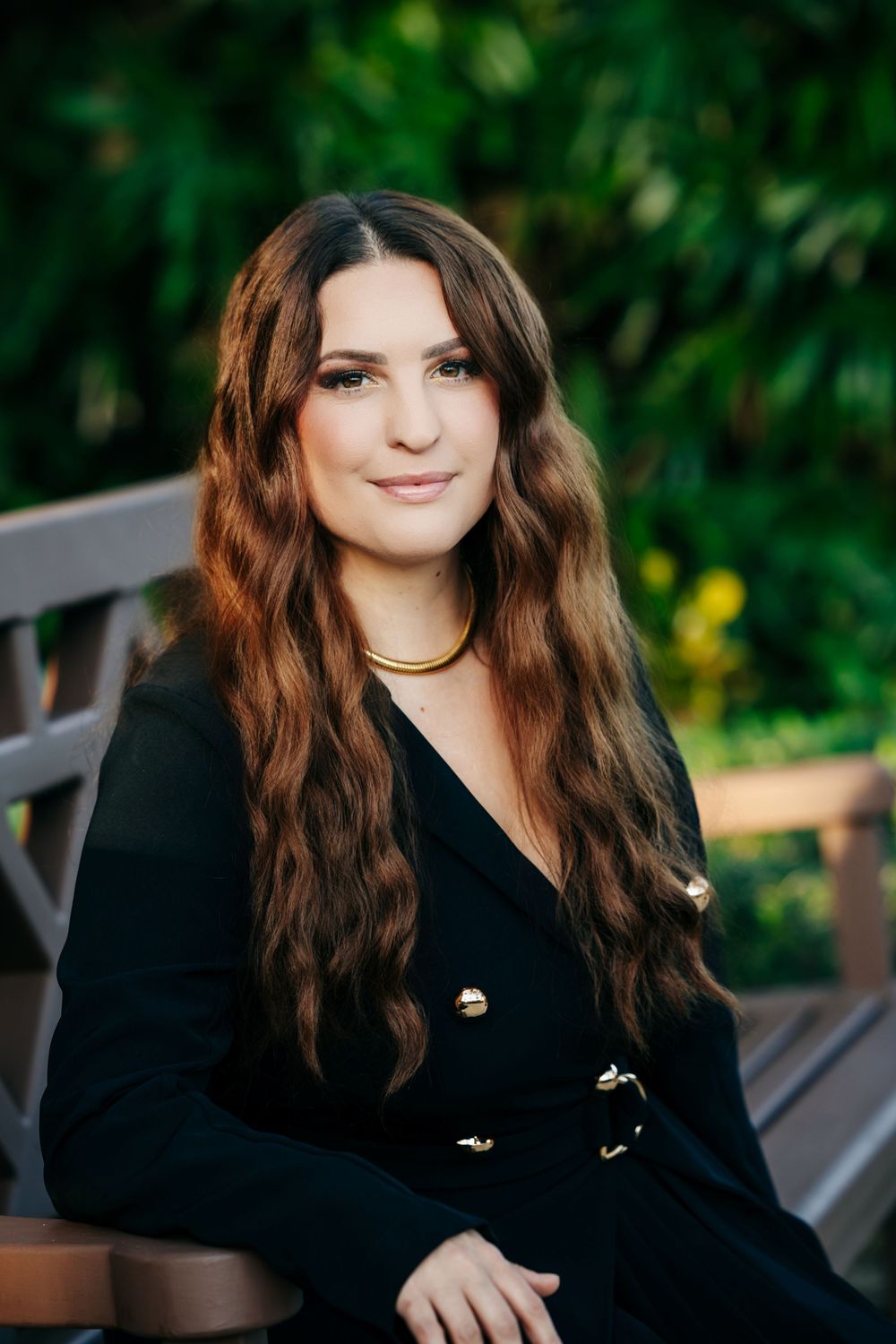 Young woman with long wavy auburn hair wearing a black blazer and gold necklace, seated outdoors on a bench.