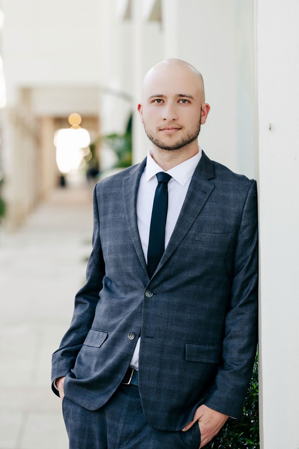 Bald businessman in gray plaid suit and black tie leaning against white pillar in modern building corridor.
