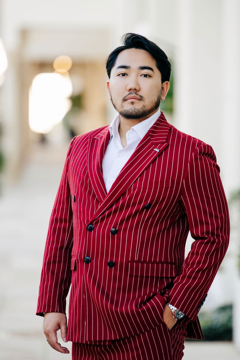 Asian man in bold red pinstripe double-breasted suit posing confidently outdoors in elegant setting.