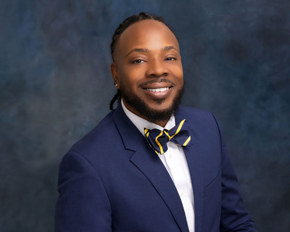 Professional man in navy blue suit and striped bow tie smiling confidently against a studio backdrop.