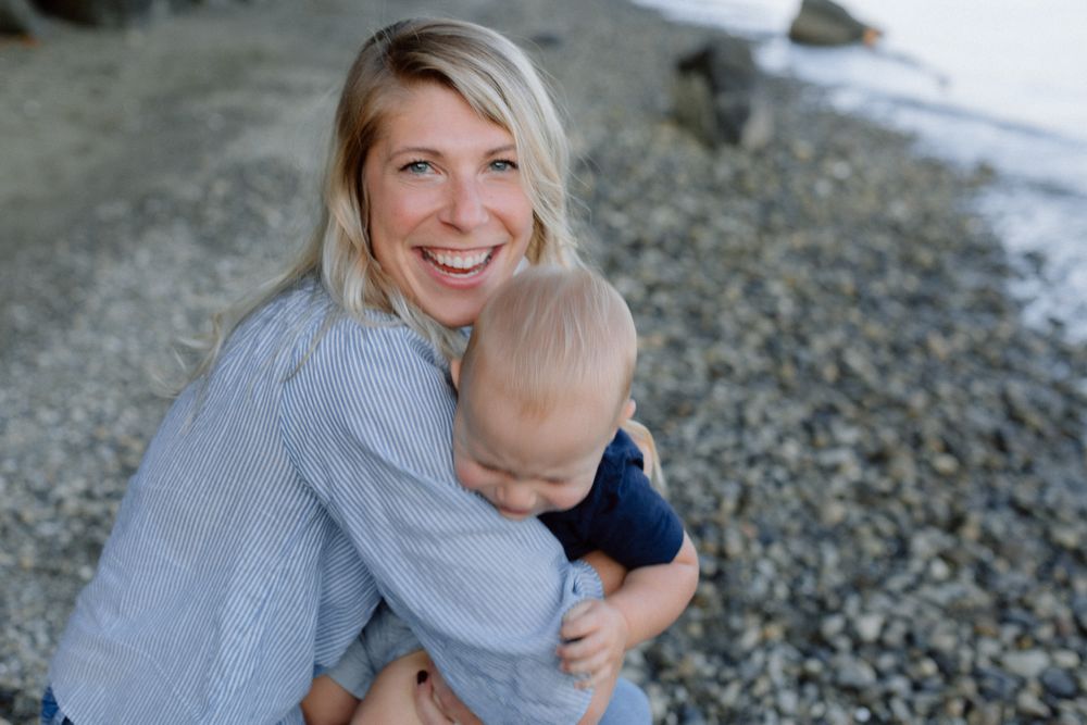 Smiling blonde woman holding a baby on a rocky beach shore.
