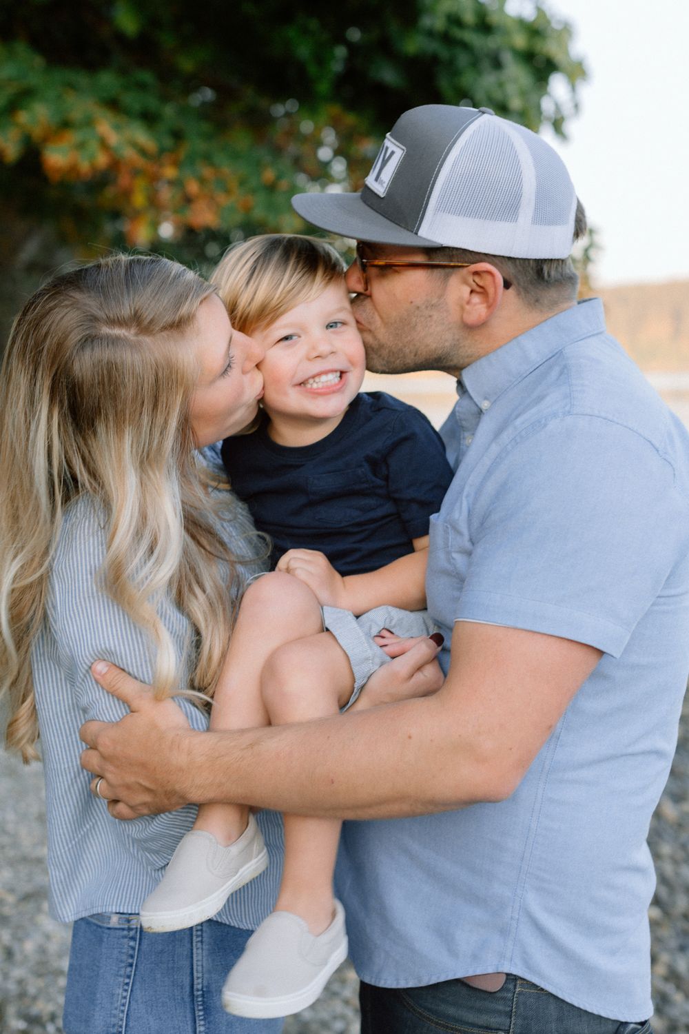 Two parents kissing their smiling toddler on the cheeks outdoors.
