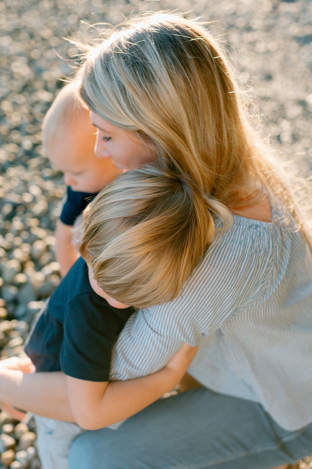 Mother tenderly hugs two young children on a rocky beach in warm golden sunlight.