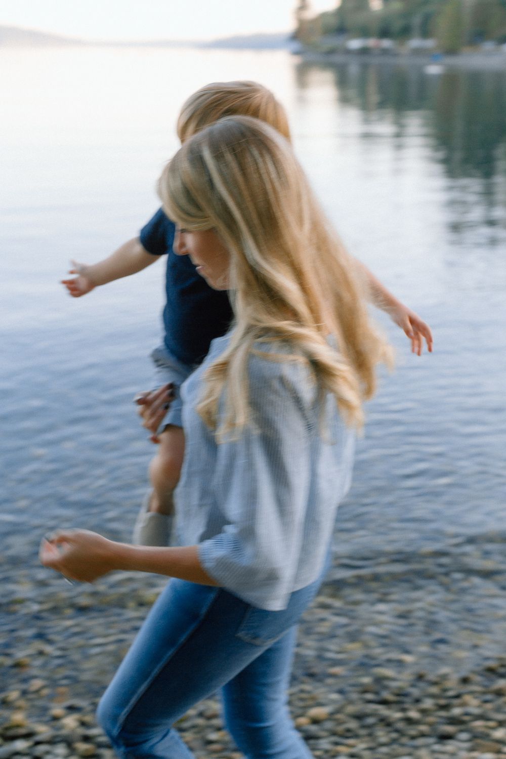 Blonde mother carrying her young child along a peaceful lakeside shoreline at dusk.