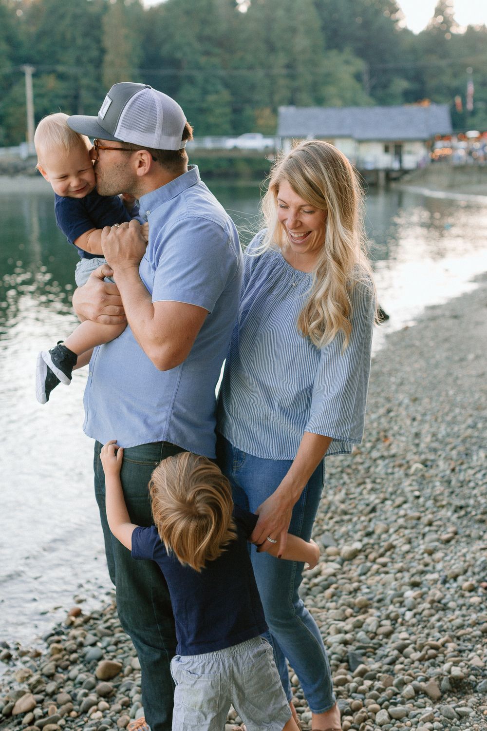 Happy family of four laughing together on a rocky beach by a calm lake at sunset.