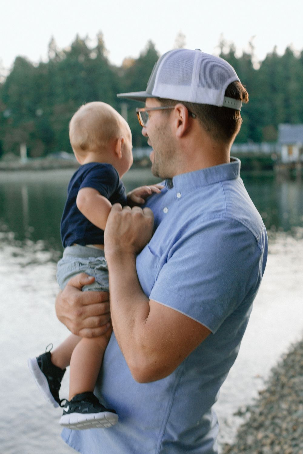 Father holding his baby son while standing by a serene lake at golden hour.