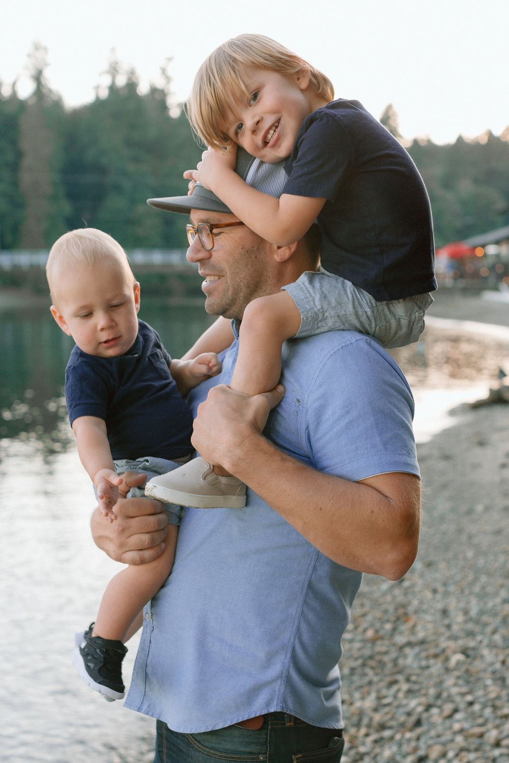 Dad carrying toddler son on shoulders while holding baby boy by a lakeside beach.