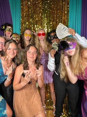 Group of party guests holding up cash and wearing festive accessories against a gold sparkly backdrop.
