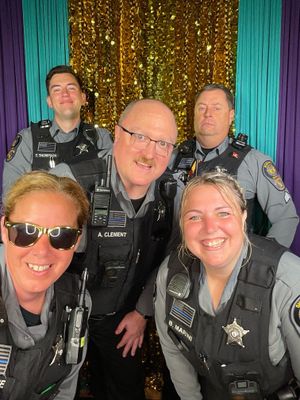 Law enforcement officers in gray uniforms pose together against a sparkly gold backdrop at an event.