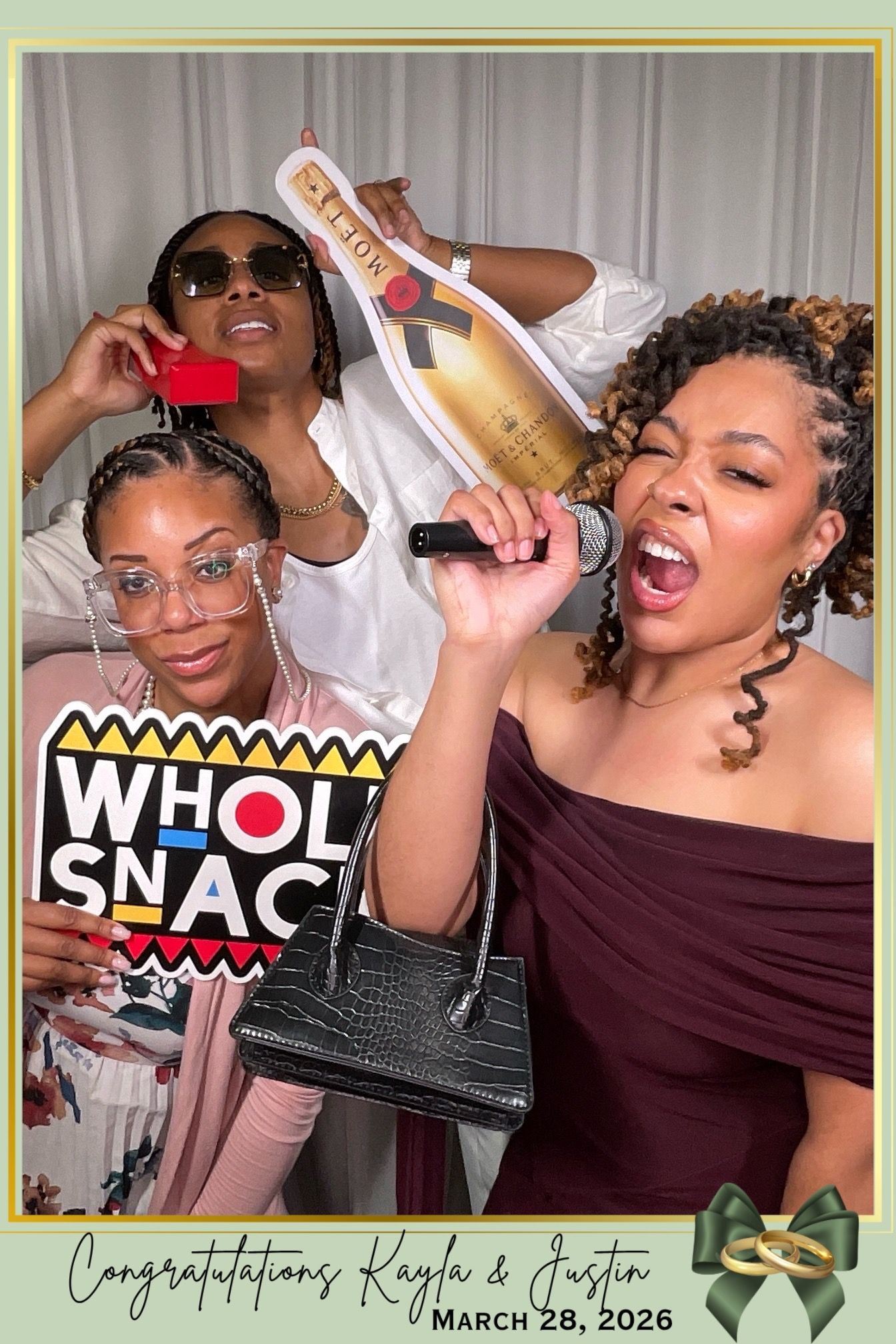 Three women pose playfully at a wedding photo booth holding props, celebrating Kayla and Justin's wedding on March 28, 2026.