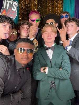 A group of people wearing sunglasses and formal attire pose together in a celebratory setting with colorful lighting.