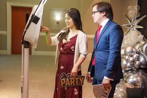 Two people use a photo booth station decorated with festive party signage and silver balloons.