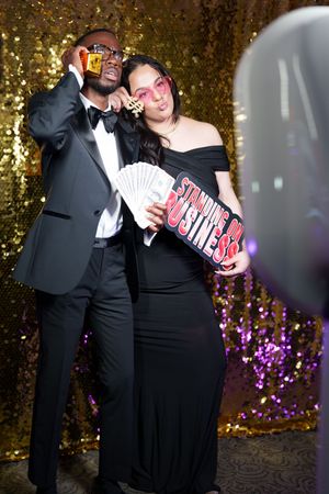 Couple in formal attire playfully posing with a box of candy against a gold backdrop with purple flowers.