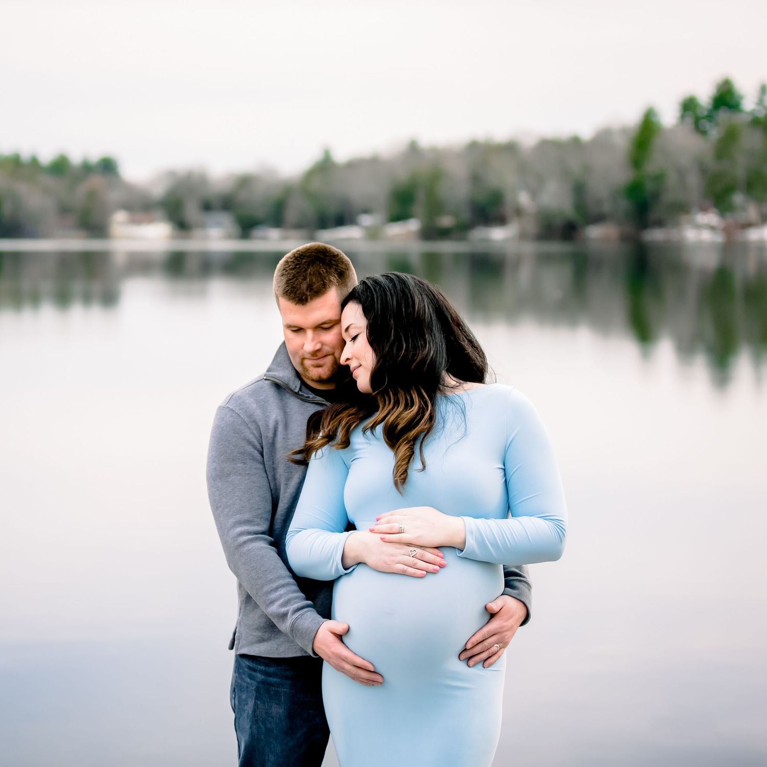 A couple in casual attire embraces by a serene lake during a maternity photoshoot on an overcast day.