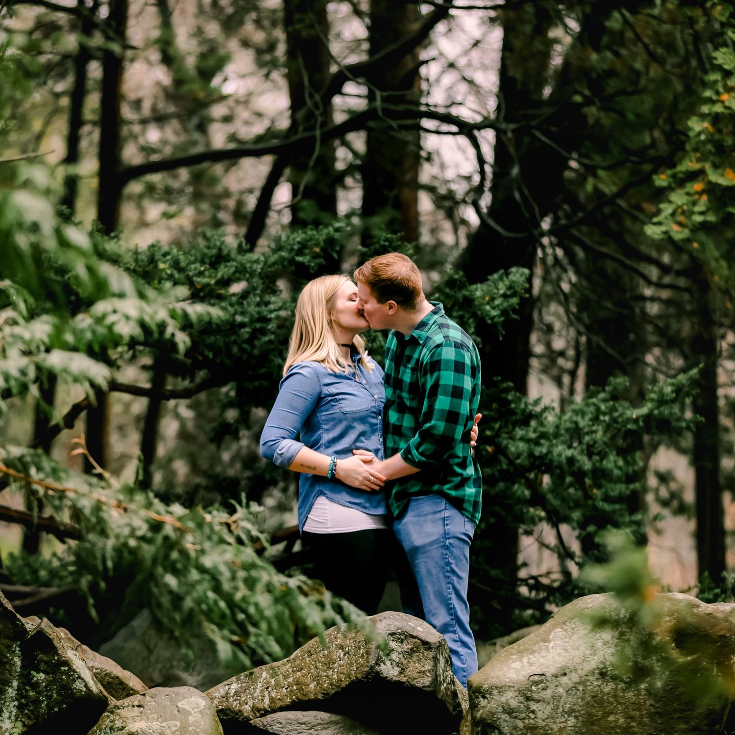 A couple in denim and plaid share an intimate moment during a pregnancy announcement sessionwhile standing on fallen logs in a misty forest.