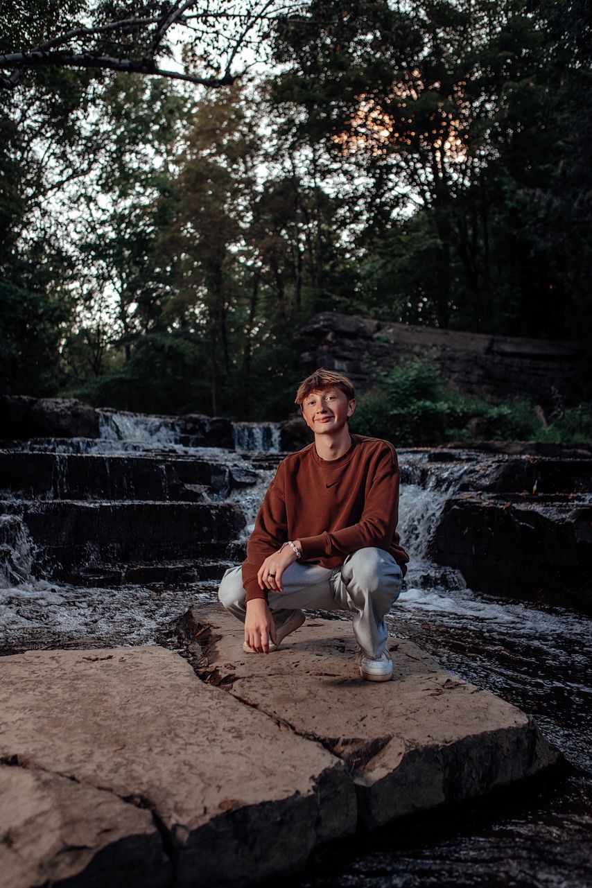 Person in brown sweater sits on rocky waterfall ledge during sunset in forest setting.