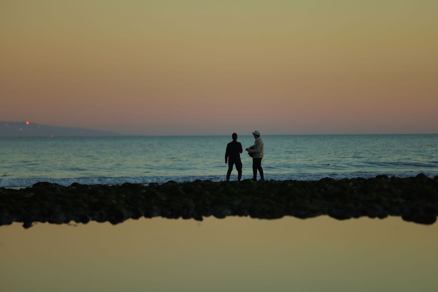 Two silhouetted figures standing on rocky shore at colorful ocean sunset.
