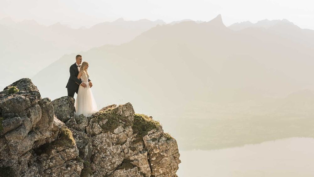 Wedding couple embracing on rocky mountain peak with misty alpine lake scenery behind them.
