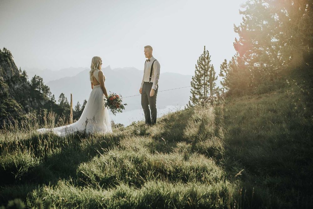 Bride and groom sharing romantic moment on sunlit mountain meadow with scenic alpine backdrop.