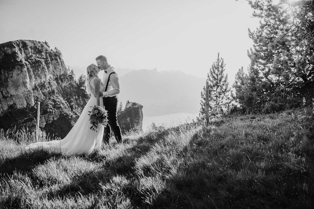 Black and white photo of wedding couple embracing on mountain hillside with rocky cliffs nearby.