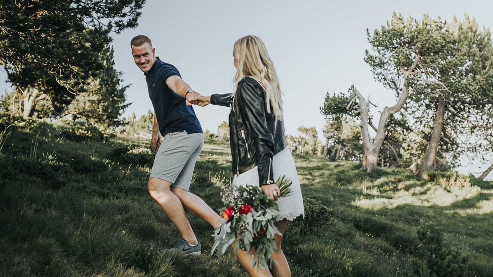 Couple holding hands walking through green hillside park with colorful bridal bouquet in hand.