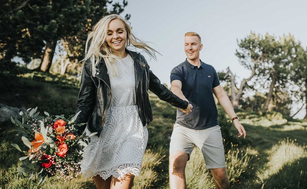 Joyful couple running through sunny green hillside, woman in white dress holding vibrant bouquet.