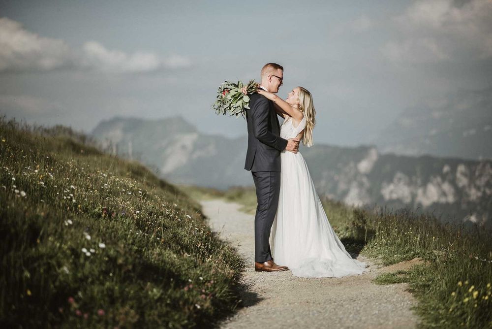 A wedding couple embracing on a scenic mountain path with alpine peaks and wildflowers behind them.