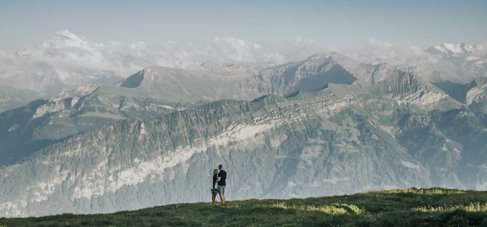 Couple standing together on a mountain summit with dramatic Alpine peaks and misty valleys stretching into the distance.
