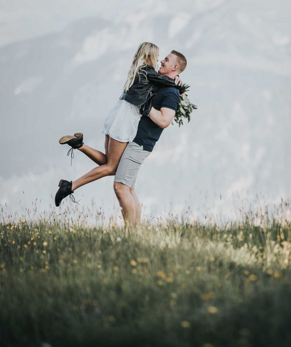 Man joyfully lifting woman in white dress in sunny alpine meadow with mountain views behind.