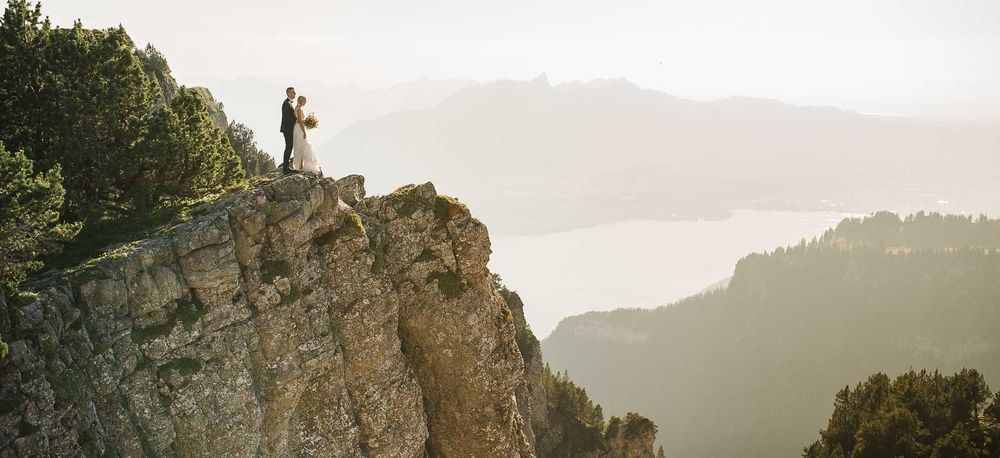 Couple eloping on dramatic rocky cliff edge with misty mountain lake views in background.