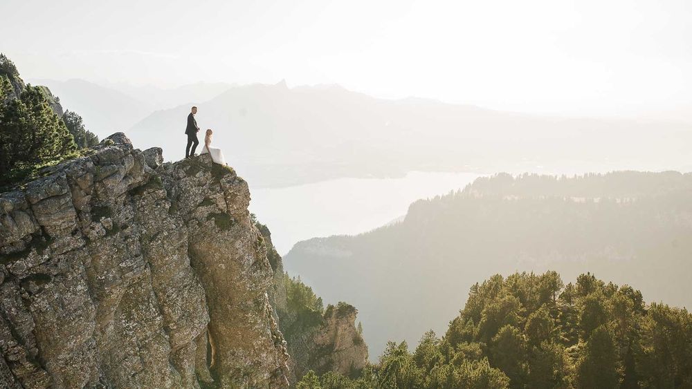 Person standing on rocky mountain summit with stunning aerial lake and valley views below.