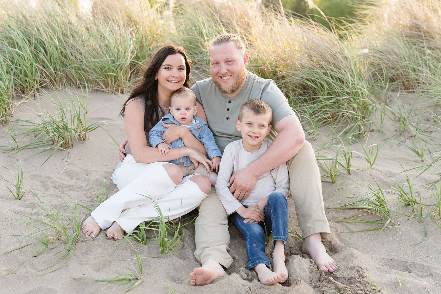 Happy family of four sitting on sandy beach dunes with tall grass, parents smiling with two young sons.