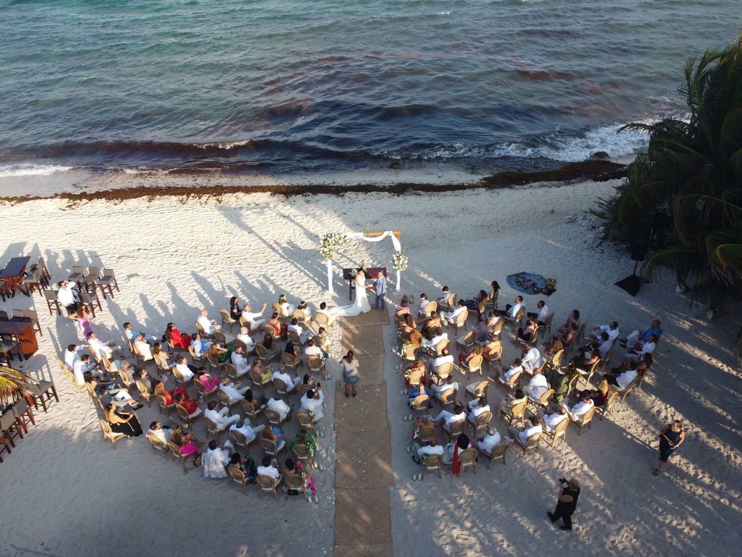 Aerial drone view of intimate beach wedding ceremony with guests seated along tropical shoreline.