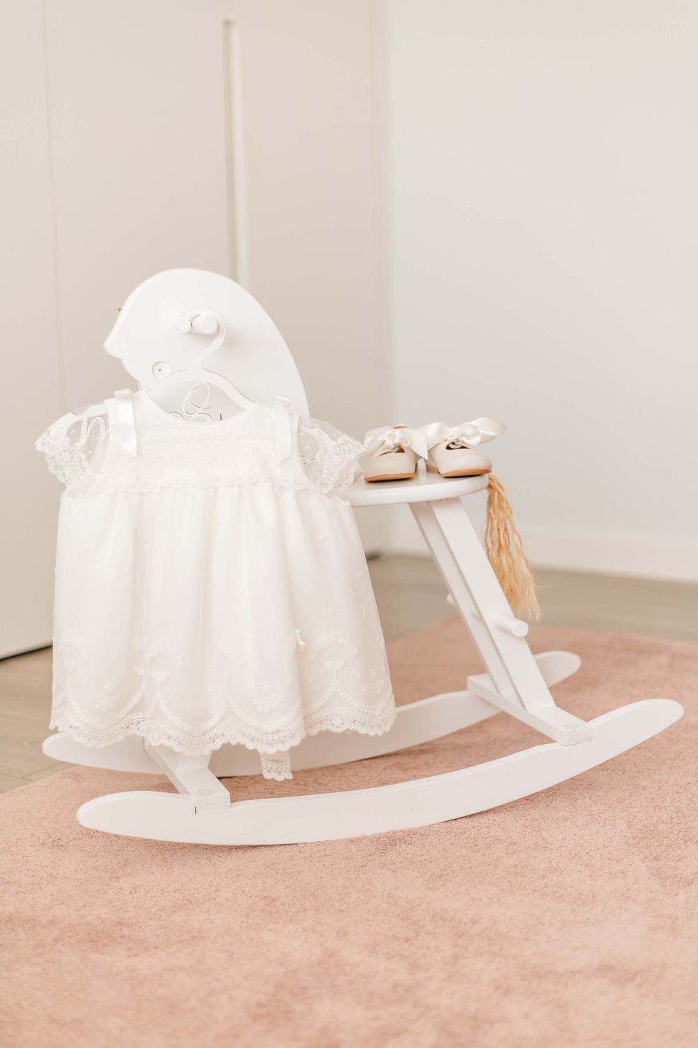 White baptismal dress displayed on rocking horse with shoes on pink carpet.