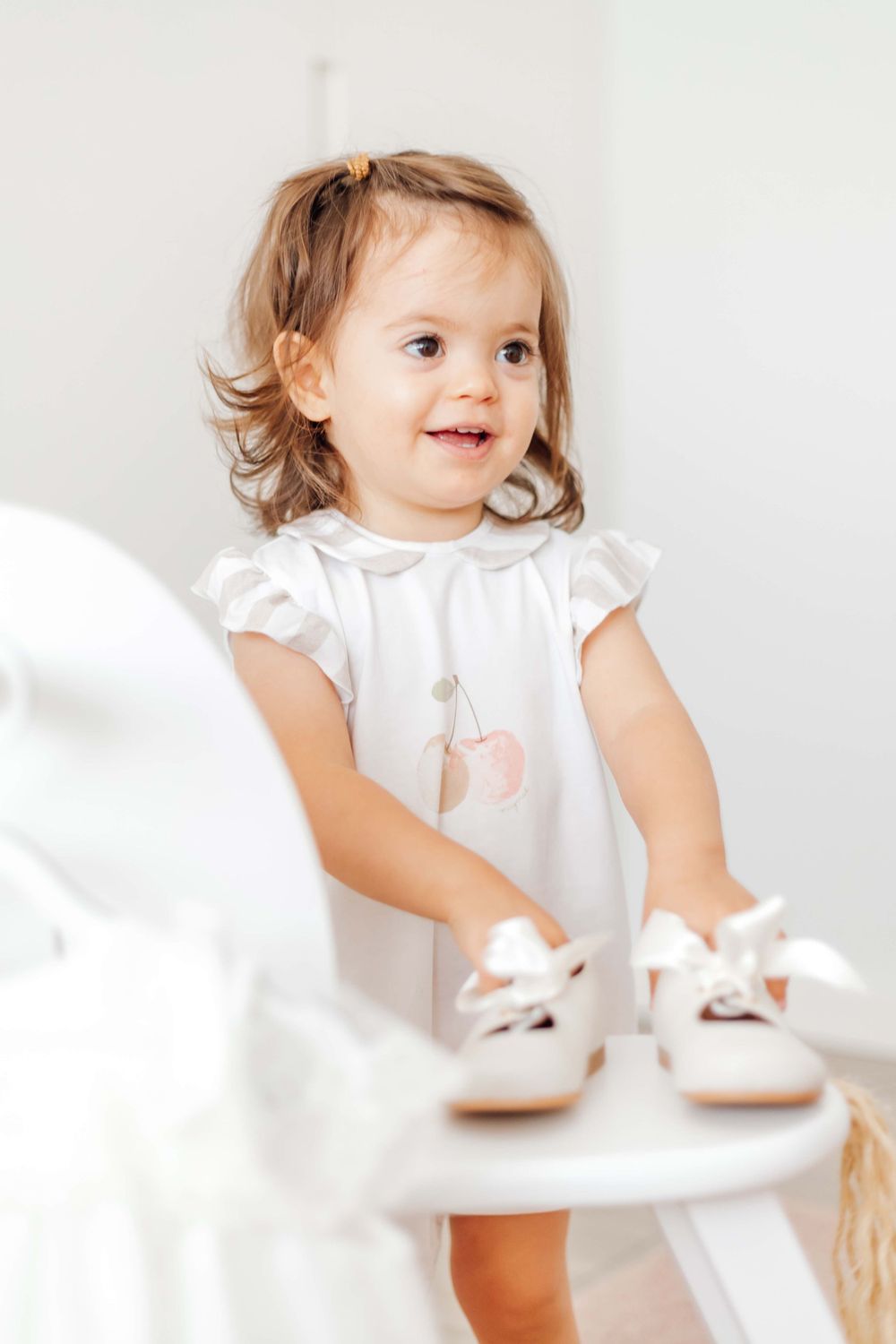 A toddler in white dress reaches towards white shoes on furniture.