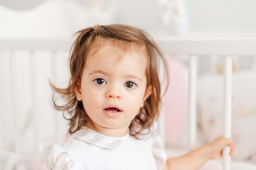 A toddler in white clothing stands near white nursery furniture.