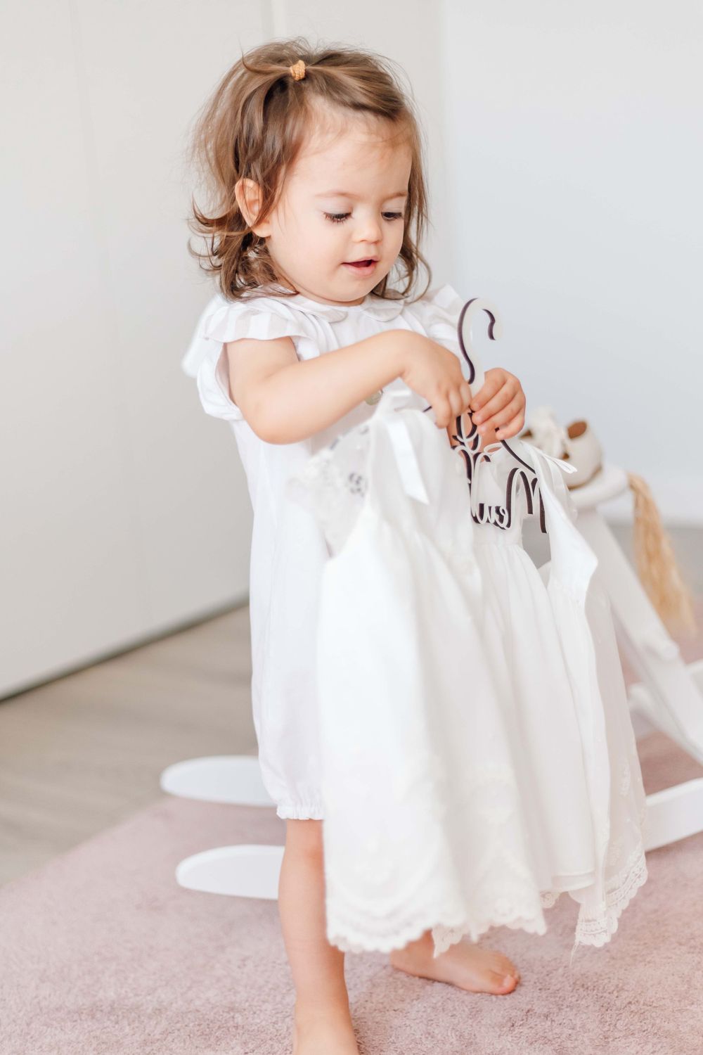 A toddler in a white dress holds a hanger while standing on a pink carpet.