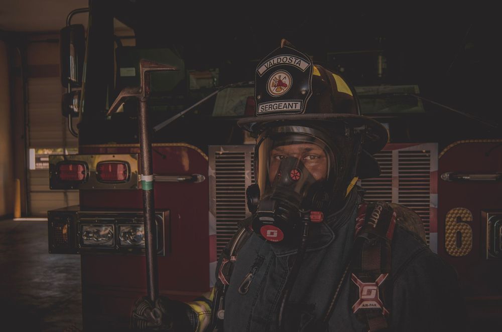 Firefighter in full gear and gas mask stands before a red fire truck in a dark dramatic setting.