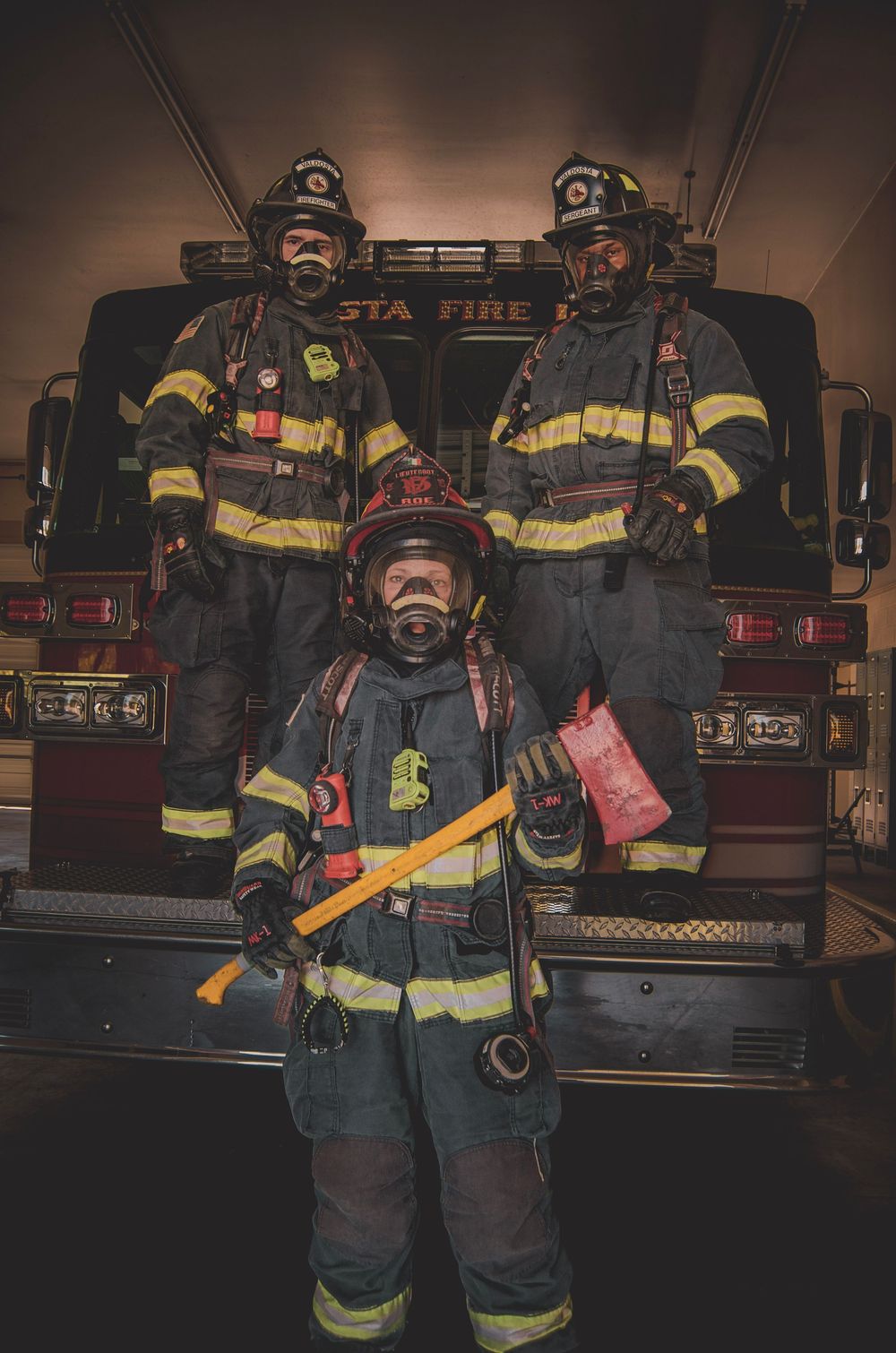 Three firefighters in full gear pose in front of a fire truck, with one holding an axe and wearing a breathing mask.
