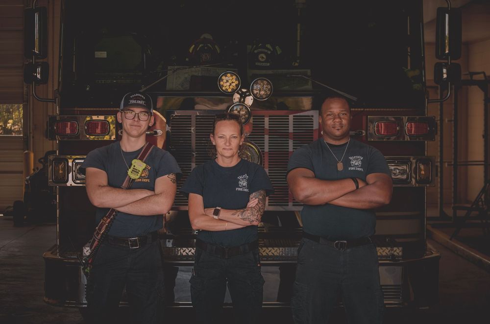 Three firefighters stand confidently with arms crossed in front of a fire truck inside a station.