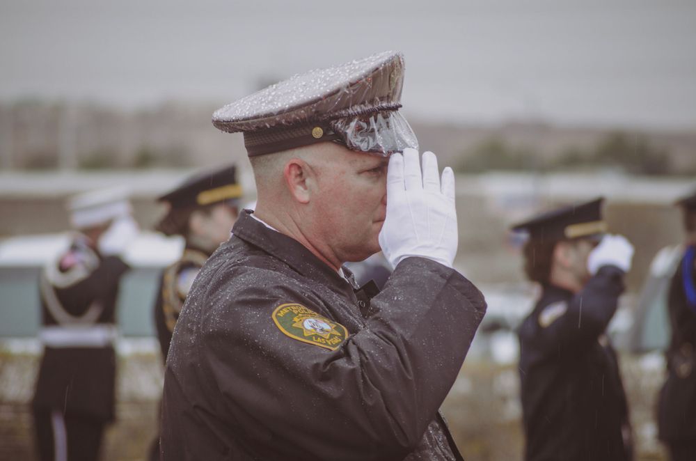 Officer in formal uniform saluting during outdoor ceremony with others visible in background.