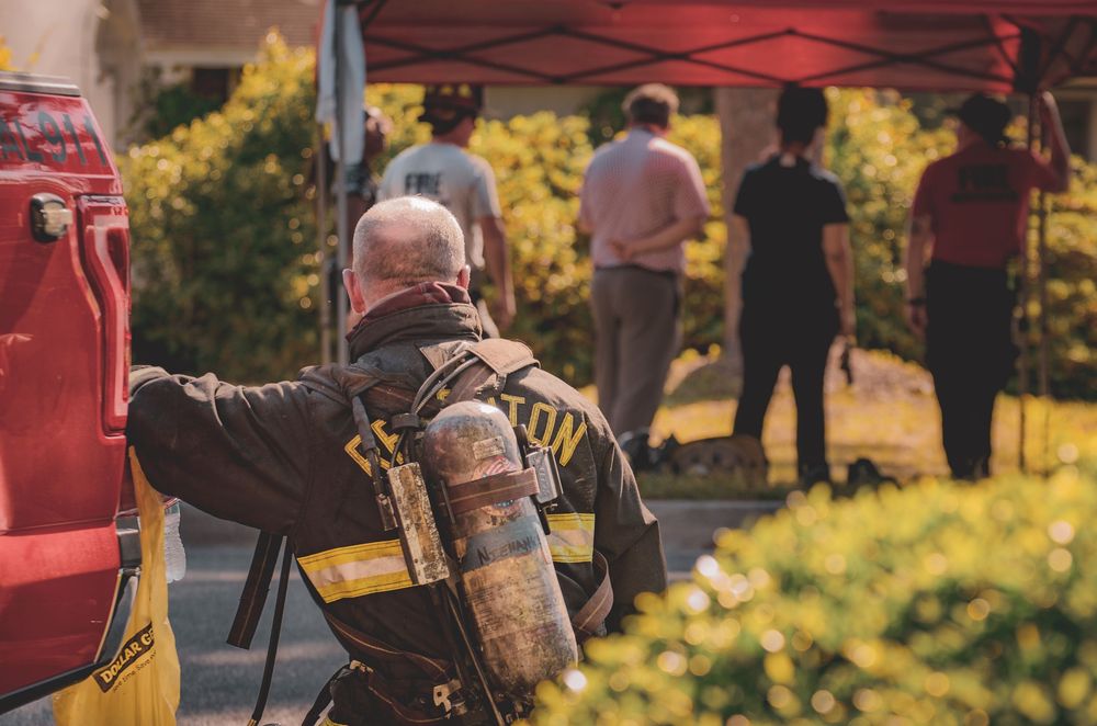Firefighter wearing SCBA tank and turnout gear stands near fire truck at outdoor community event.