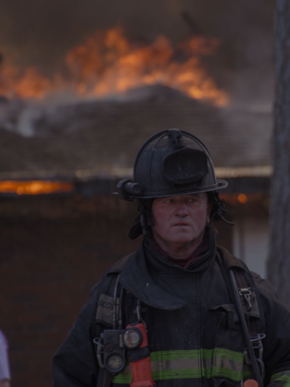 Firefighter in black helmet and gear stands before a blazing structure fire, looking directly at the camera.