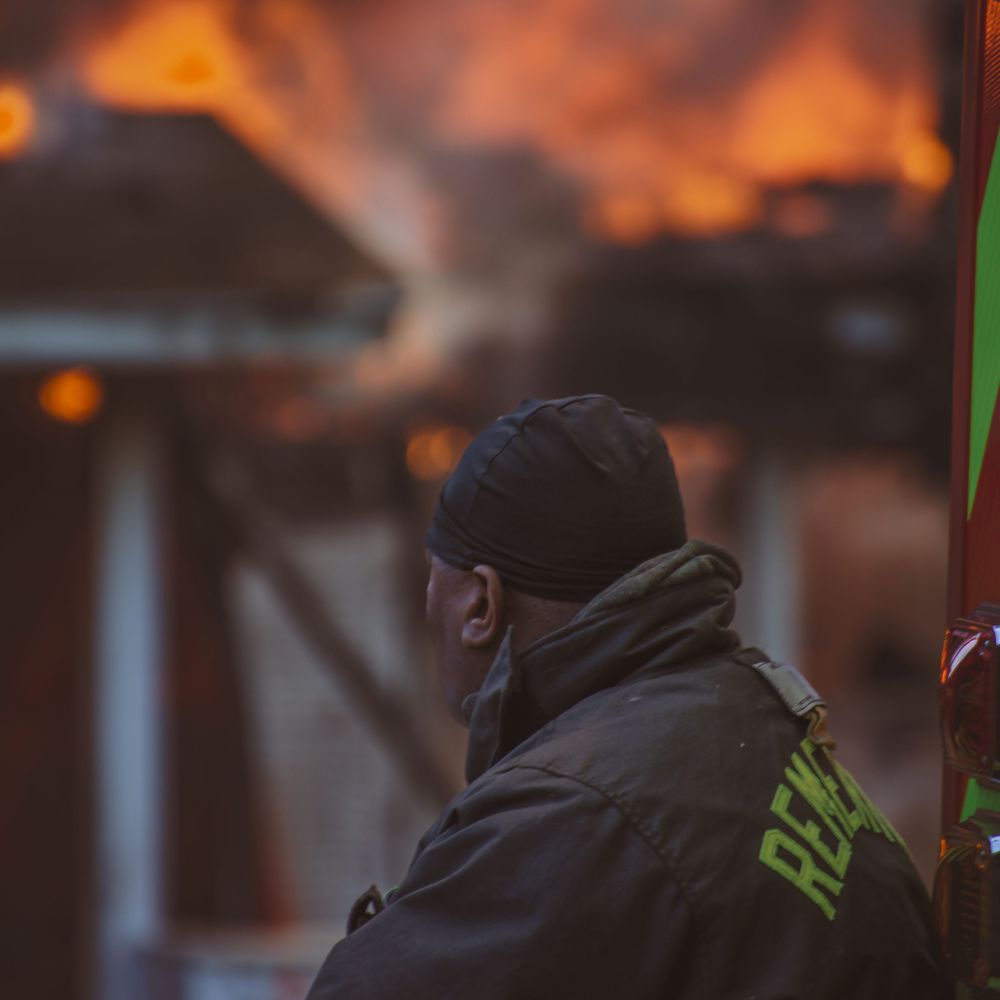 Firefighter in dark jacket with RESCUE text watches a building engulfed in flames during an emergency response.