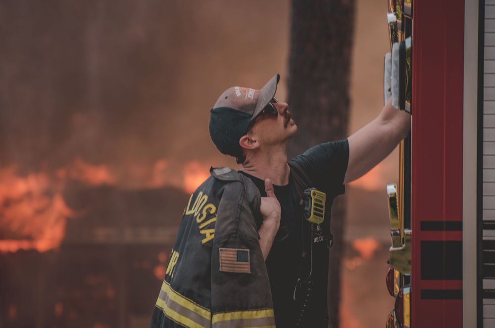 Firefighter in gear and cap reaches up to a fire truck amid intense flames and smoke in the background.
