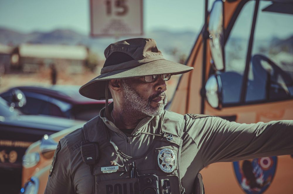 Law enforcement officer in wide-brimmed hat and tactical vest standing next to vehicle in bright sunlight.