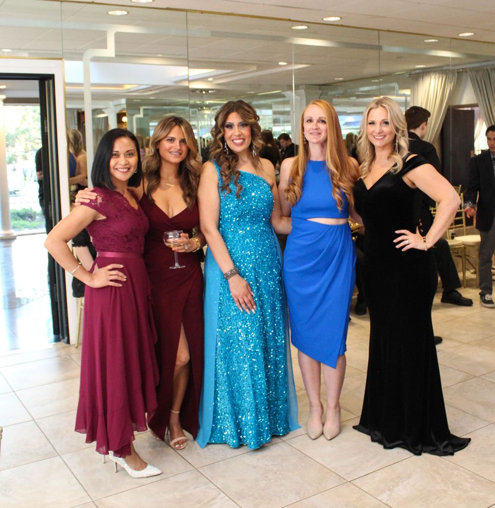 Five women in formal dresses pose together in an elegant hallway setting.