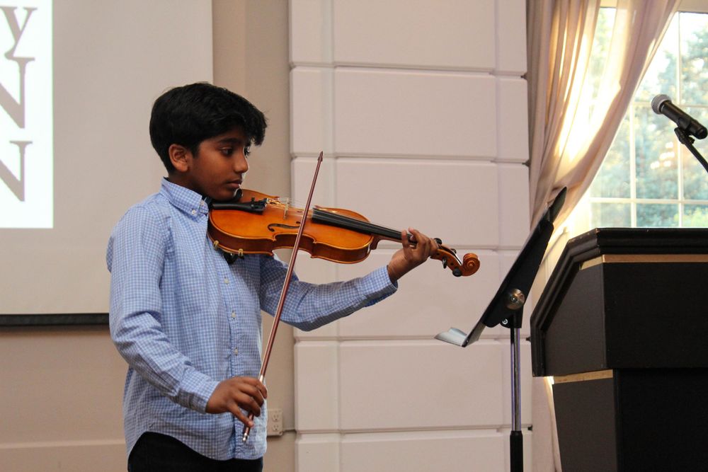 A young musician in a light blue shirt performs on violin with sheet music stand.