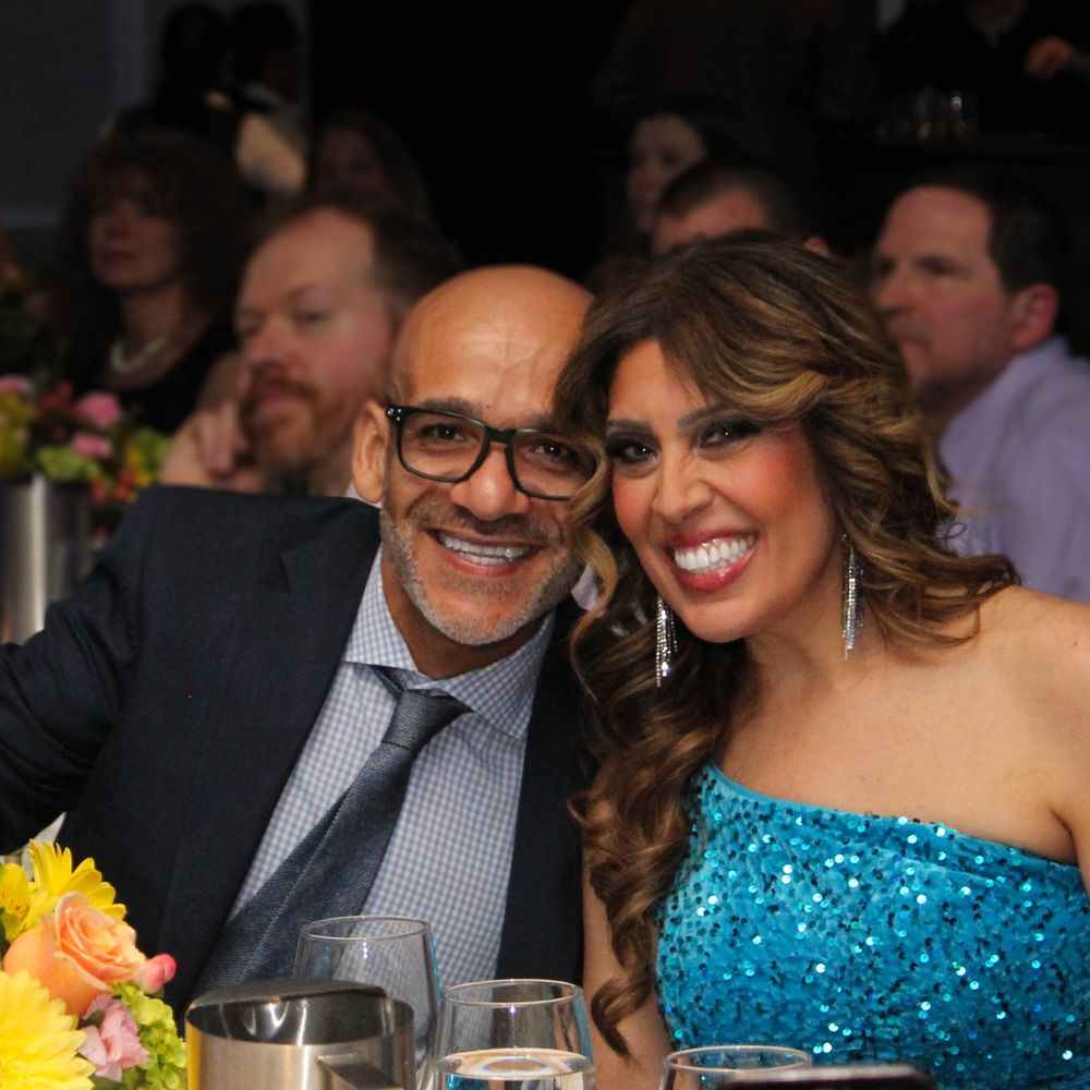 A couple in formalwear smiles at a dinner table decorated with yellow flowers.