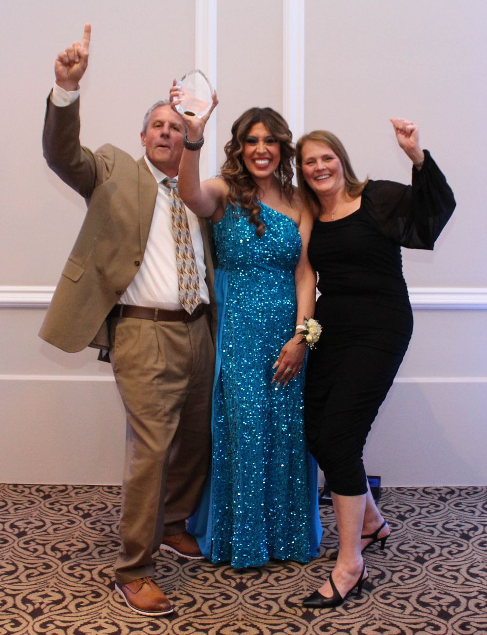 Three partygoers celebrate with raised arms, featuring a sparkly blue sequined evening gown at a formal event.